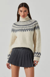 A woman is wearing a cream-colored, chunky knit turtleneck sweater with a gray geometric pattern around the yoke, cuffs, and hem. She pairs it with a high-waisted, black pleated mini skirt featuring thin vertical stripes.